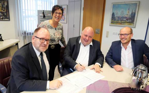 Drei Männer sitzen an einem Tisch in einem hellen Büro, eine Frau steht dahinter. Der mann in der Mitte unterzeichnet einen Vertrag, alle schauen in die Kamera.