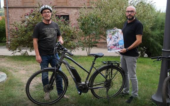 Zwei Herren hinter einem Fahrrad. Der linke Herr trägt einen Helm und hält den Lenker des Fahrrads fest. Der rechte Herr zeigt das Plakat zum STADTRADELN.