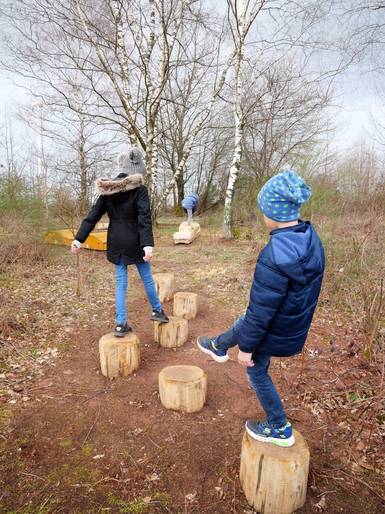 Drei Kinder in Winterkleidung balancieren auf Holzpflöcken.